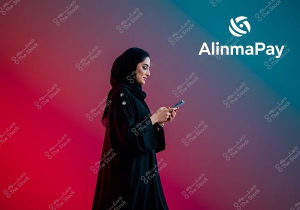 An image of a woman wearing a black abaya standing against a colorful background blending blue and pink. She is holding a mobile phone, looking at it with a slight smile. The \"Alinma Pay\" logo is visible in the top right corner of the image.