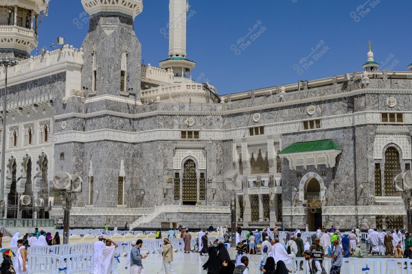 An image of a group of people walking around the Masjid al-Haram in Mecca, Saudi Arabia. The image shows part of the Kaaba and the intricately decorated marble walls of the mosque with Islamic motifs. People are wearing white Ihram garments, and others are in diverse clothing representing various cultures.