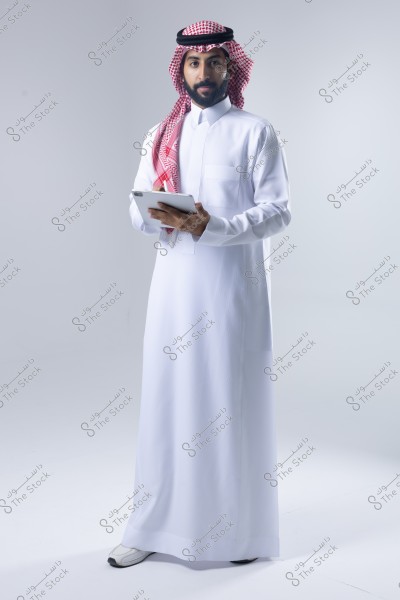صورة لرجل يرتدي الثوب التقليدي السعودي الأبيض والغترة الحمراء والبيضاء على رأسه, ويمسك بجهاز لوحي. يقف الرجل في استوديو بإضاءة ساطعة وخلفية بيضاء.