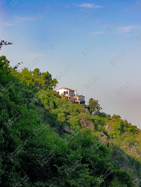 An image of a house situated on the side of a hill covered in dense green foliage. The house is at the top, appearing white with a red roof, surrounded by trees and greenery. The sky is clear blue with a few scattered clouds.