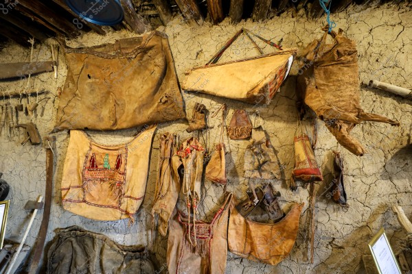 An image of a mud wall displaying traditional tools and objects made from leather and natural materials. Large and small leather bags with various colors and patterns are hanging on the wall. The flooring and interior design suggest an old cultural heritage.