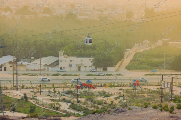 تظهر الصورة مشهداً لمدينة بهضبة مرتفعة حيث يمكن رؤية تلفريك معلق في الهواء يمر فوق منطقة خضراء مليئة بالأشجار والنخيل. في الأمام, يظهر متنزه للأطفال يحتوي على ألعاب ملونة, مثل الزلاجات والأرجوحات. يمكن أيضاً رؤية شارع مزدحم بعدة سيارات, ومباني متعددة الطوابق في الخلفية.