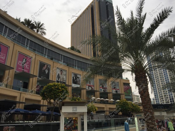 A view of a shopping mall facade in a modern city, featuring a high-rise building with skyscrapers in the background. Palm trees and part of the walkway are prominent in the scene, along with large advertisement banners for well-known brands. The weather is cloudy, resembling the atmosphere of Dubai in the United Arab Emirates.