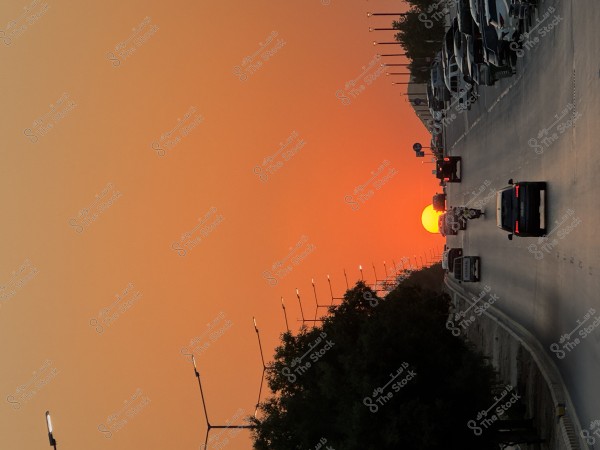 A busy road with cars at sunset, where the sun \r\n\r\nappears large and red on the horizon, with the sky glowing orange. Trees are visible on the left side of the road and streetlights are illuminated.