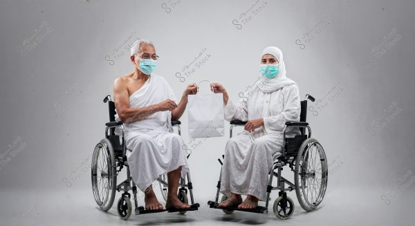 The image shows a man and a woman sitting in wheelchairs in a studio with a gray background. The man is wearing white Ihram clothing, and the woman is wearing a hijab and modest white clothing. Both are wearing blue medical masks. They are jointly holding a white shopping bag between them. The scene may suggest preparation for Hajj or Umrah.