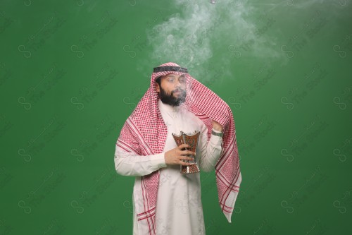 رجل سعودي يقف على خلفية خضراء يرتدي الثوب و الشماغ، يقوم بعمل حركات بيديه ، يبخر نفسه بالطيب و العود، احتفالات العيد