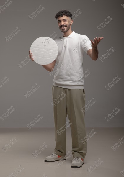 A portrait of a man standing in a photography studio. The man is wearing a white polo shirt, beige trousers, and white sneakers. He holds a round white disk in his left hand and appears to be smiling. The background is a plain gray.