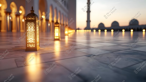 Illuminated lanterns inside a mosque, placed on a wide marble courtyard at sunset. The lanterns are lit in harmony with the lights lining the courtyard, and domes are visible in the background.