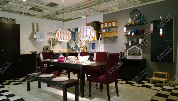 A modern dining room in a furniture store. It features a black dining table surrounded by chairs with purple upholstery. Above the table, there are two pendant lamps with a contemporary design. The walls are decorated with shelves holding plates, glasses, and decorative vases. The floor has a black and white checkered pattern and a light-colored rug.