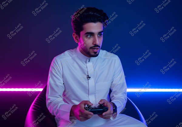 A portrait of a young man wearing a traditional white Saudi thobe, sitting on a comfortable chair and holding a video game controller in his hands. The background lighting consists of purple and blue, adding a modern and warm atmosphere to the image.