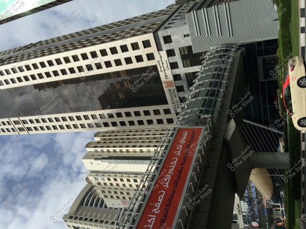 The image features modern skyscrapers in a city filled with tall buildings. Prominently in the foreground is the \"Millennium Plaza Hotel\" building with a glass pedestrian bridge spanning the street. An orange advertisement is displayed on the bridge. Cars are seen below, reflecting the vibrant city life. The sky is blue with some white clouds.