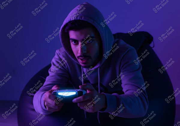 A person sitting on a beanbag, wearing a hooded sweatshirt and playing with a game controller. The purple and blue lighting adds an exciting atmosphere. The person appears focused and immersed in the game.