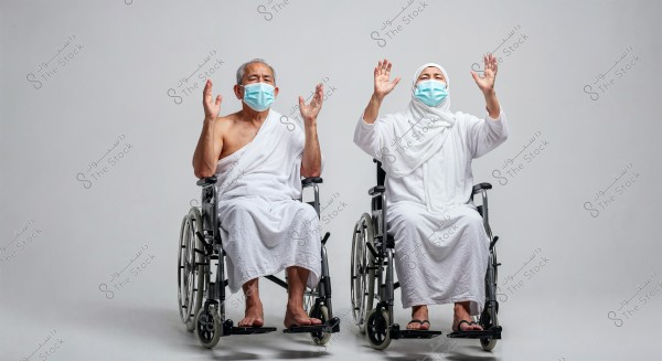 An elderly man and woman sitting in wheelchairs against a white background. Both are wearing traditional Ihram clothing, which is a white garment worn for Umrah or Hajj. The man is wearing a single piece of cloth draped over his upper body and a white lower garment, while the woman is dressed in a long white robe with a headscarf. Both have their hands raised as if in prayer. They are also wearing blue protective masks.