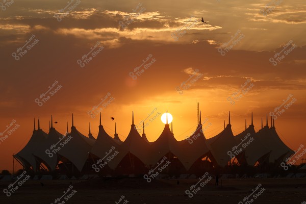 Image of the sunset behind King Fahd International Stadium in Riyadh, Saudi Arabia. The structure features tent-like conical forms, with the sky illuminated in warm orange and purple hues. Birds are flying across the sky, adding to the visual grandeur of the scene.