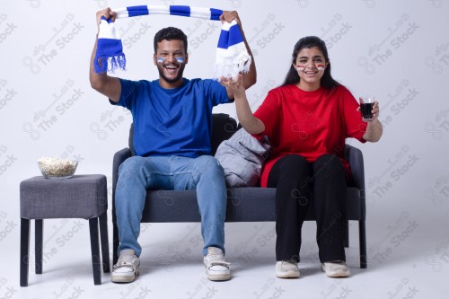 Portrait of a Saudi young man and woman watching a match and cheering their favorite teams cheerfully while having a snack on a blue background, the World Cup.