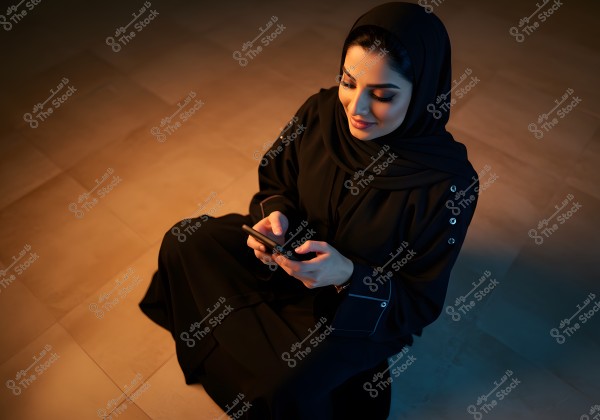 A woman sitting on the floor, wearing a black abaya and hijab, looking at a smartphone in her hands. The lighting is soft and warm on the left side of the image, creating a cozy and calm atmosphere.