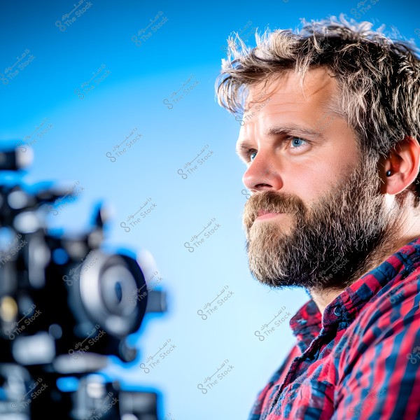 A photo of a bearded man with short blonde hair wearing a red and blue plaid shirt. The man is shown in profile looking forward, with a large camera visible in the background. The background is light blue.