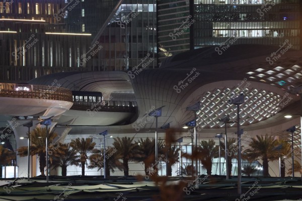 A nighttime urban scene featuring a modern building with an impressive architectural design, including curves and innovative patterns. In the foreground, palm trees are lined alongside light poles, with skyscrapers showcasing glowing nighttime lights in the background.