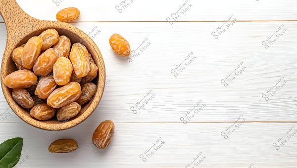 An image showing a collection of golden dates inside a wooden bowl on a white wooden background. The dates appear glossy and smooth, with some scattered outside the bowl. A green leaf is visible in the lower left corner.