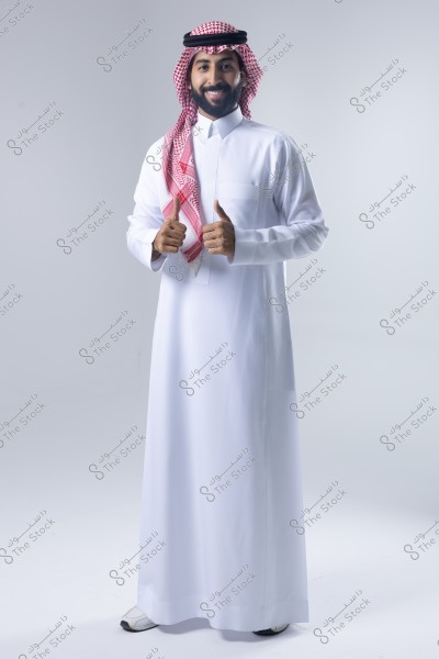 A portrait of a man standing and smiling, wearing a traditional white thobe, a red and white ghutra, and a black agal. The man is in a positive pose with both thumbs up. The traditional clothing suggests he is from the Arabian Gulf region, likely Saudi Arabia.
