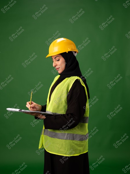 Image of a woman wearing a yellow reflective vest and a yellow safety helmet, holding a clipboard and looking at it. She is dressed in a black abaya and black hijab. The background is green.