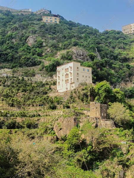 منظر طبيعي لجبل مغطى بالخضرة والنباتات الكثيفة في منطقة جبلية. تظهر عدة مبانٍ ذات تصميم معماري تقليدي على المدرجات الجبلية؛ أحدها مبني من الحجر, والآخر بلون فاتح بارز. السماء صافية وتشير إلى يوم مشمس.