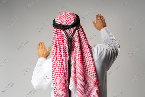 Image of a man in traditional Arab attire from the back. The man is wearing a white thobe and a red keffiyeh with a black agal. He appears to be raising his hands in a gesture of prayer or greeting. The background is light grey.