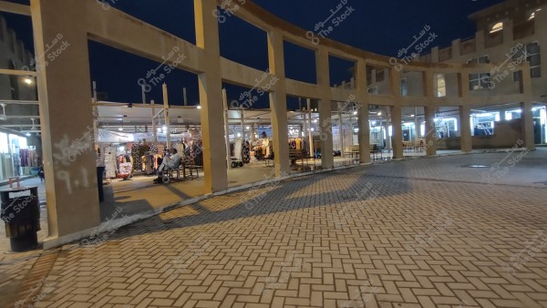 Nighttime image of an outdoor market with an architectural structure featuring open columns forming rectangles, and a tiled ground. Illuminated clothing shops are visible in the background, displaying various fabrics and garments, with a person seated on a bench. On the left side, a trash bin and a few shoppers are seen.