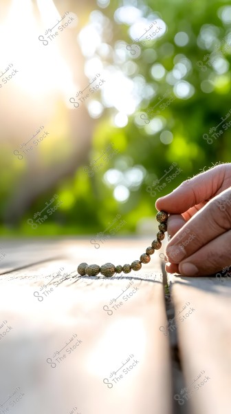 A hand holding a set of brown prayer beads on a wooden surface under strong sunlight. The background is blurred, showing green hues from plants, suggesting an outdoor area or garden.