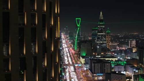 منظر ليلي لمدينة الرياض يُظهر برجي المملكة والفيصلية مضاءين بالأضواء الخضراء, مع حركة مرورية كثيفة على الطريق الرئيسي بين المباني الشاهقة.\r\n\r\n-