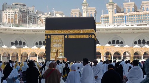 مشاهد من الفيديو تعرض الكعبة في مكة المكرمة أثناء الطواف, حيث يظهر العديد من الحجاج يرتدون ملابس الإحرام ويمارسون الشعائر الدينية في ساحة الحرم المكي الشريف, تحت سماء زرقاء ومباني برجية في الخلفية.