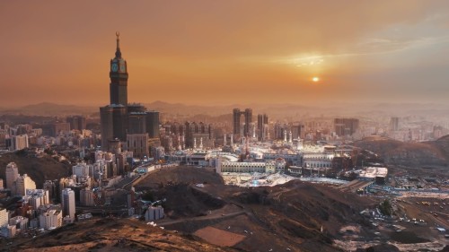 مشهد جوي لمدينة مكة المكرمة يظهر فيها الحرم المكي الشريف وبرج الساعة في وقت الشروق, مع سماء تتخذ لوناً دافئاً.