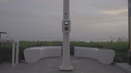 The video shows a scene of an advanced electric charging station equipped with modern technology by the seaside. A person approaches the station in a calm atmosphere surrounded by lush greenery and flowers. The video highlights the development of clean energy infrastructure.