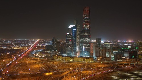 منظر جوي ليلي يُظهر معالم أفق مدينة الرياض بإضاءة ليلية براقة لناطحات السحاب والشوارع التي تمتد طويلاً بإضاءات السيارات. تظهر المباني المعمارية الحديثة والتقاطعات المزدحمة بالسيارات في قلب المدينة متعددة الأضواء.