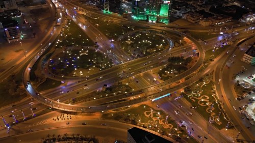 فيديو لقطات جوية لطريق دائري مضاء في مدينة الرياض ليلاً, تُظهر تصميمات معمارية مميزة وتنسيق حدائق مضاءة بأضواء ملونة, مع حركة مرور نشطة تشير إلى حياة المدينة الليلية الديناميكية.