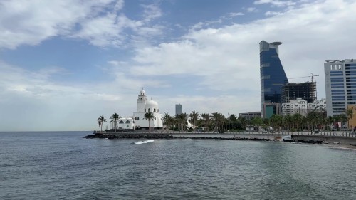 A scenic view showcasing a distinctive white mosque by the sea, surrounded by palm trees and modern buildings under a sunny blue sky.\r\n\r\n-