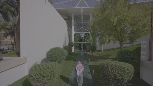 تظهر اللقطات شخصًا يرتدي الزي التقليدي السعودي يسير في ممر حديث محاط بالأشجار والنباتات الخضراء أمام مبنى ذو واجهات زجاجية وبنية هندسية حديثة.