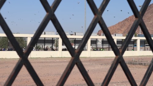 لقطة تُظهر مقبرة أُحد بالمدينة المنورة من خلف سياج معدني تحتوي على جدار ذو شبكة حديدية, وتظهر جبال في الخلفية.