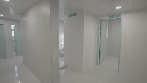 The video showcases a clean hallway in a public facility, featuring multiple room doors, including one designated for accessibility needs. The hallway has brightly lit walls and ceiling, with light reflecting on the doors in a calm, sea-like color. There is an area dedicated to ablution with taps and seats.