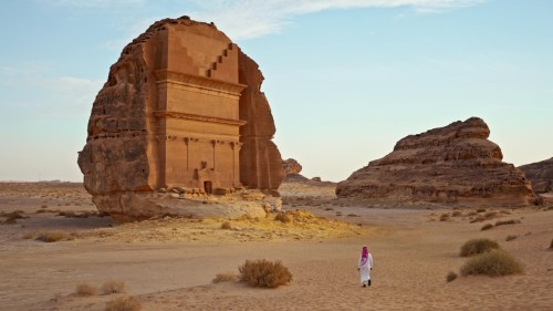 فيديو يعرض مشاهد من موقع الحجر التاريخي في العلا, المملكة العربية السعودية, يظهر فيه شخص يرتدي الزي التقليدي السعودي يمشي بالقرب من القصر الروماني المحفور في الصخر وسط الصحراء الشاسعة تحت سماء مشرقة.