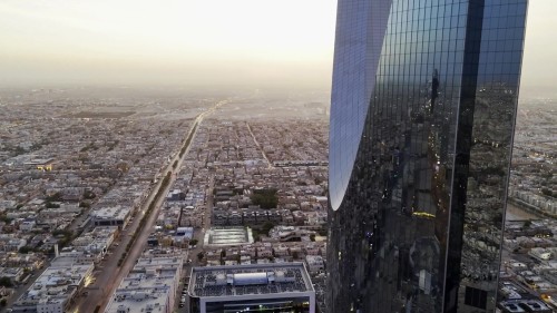 مشهد جوي لمدينة الرياض عند الغروب, يظهر فيه برج بارز ومناظر المدينة الواسعة والمباني المحيطة به. يمكن رؤية شوارع المدينة تمتد بالأفق مع السيارات المتحركة وأضواء المباني المشعة.