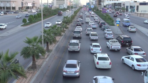 فيديو يعرض مشهداً مزدحماً لحركة المرور في شارع رئيسي في مدينة الرياض. توجد العديد من السيارات والمركبات المختلفة تسير في كلا الاتجاهين, مع وجود أشجار النخيل على الجانبين مبعثرة على جانبي الطريق. يظهر المباني التجارية والمحلات في الخلفية.