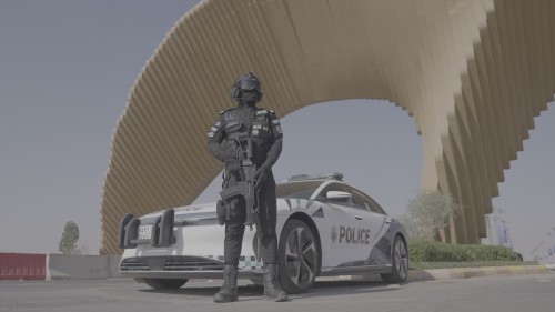 يظهر الفيديو ضابط شرطة بزي مدجج بالسلاح يقف بجانب سيارة شرطة تحت هيكل معماري حديث في بيئة حضرية, مع وجود سيارة شرطة أخرى في الخلفية. يُبرز الفيديو الإعداد الأمني والتواجد الشرطي المتقدم.\r\n\r\n-