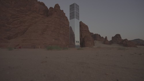 يعرض الفيديو صرحًا معدنيًا طويلًا في وسط صحراء صخرية، يعكس صورًا للبيئة المحيطة به أثناء شروق الشمس أو غروبها.