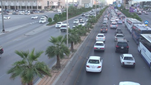 مشهد شارع مزدحم في مدينة الرياض خلال وقت الذروة النهارية, حيث تظهر السيارات المتوقفة في طابور طويل. يوجد حزام أخضر من الأشجار يفصل بين مساري الشارع, بينما يتدفق المرور بالاتجاه الآخر بشكل أكثر سلاسة.