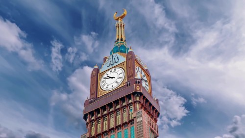 فيديو يُظهر برج الساعة في مكة المكرمة تحت سماء زرقاء صافية مع سحب بيضاء. يُظهر تفاصيل التصميم المعماري والزخارف الإسلامية على البرج الذي يُعتبر جزءاً من أبراج البيت.