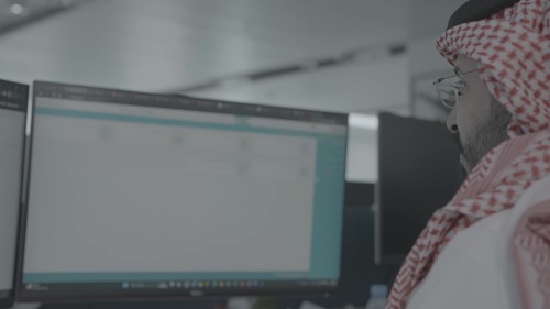 A man in an office wearing traditional Gulf attire, focuses on a computer screen while performing office work. The screen displays a software or electronic system used for daily tasks.\r\n\r\n-