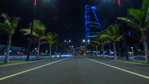 The video shows Jeddah corniche Street at night, flanked by palm trees and buildings illuminated with bright blue lights, capturing the trailing lights of fast-moving traffic.