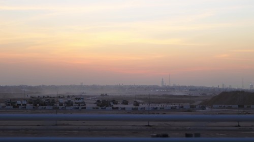 Sunset views over a city, showcasing the distant city skyline with warm gradient colors in the sky, and silhouettes of buildings and sand dunes in the foreground.\r\n\r\n-