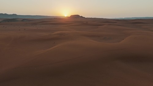 لقطات فيديو جوية لغروب الشمس في صحراء شاسعة, تظهر الكثبان الرملية ذات اللون البرتقالي والجبال في الأفق. الضوء الذهبي للشمس يضفي جواً هادئاً وساحراً على المنظر الطبيعي.\r\n\r\n-